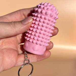 Può includere: Un portachiavi cilindrico rosa chiaro caratterizzato da una superficie ruvida coperta di piccole punte. L'oggetto presenta una base liscia ed è collegato a un anello in metallo color argento tramite una catenella, con un design tattile e spinoso.