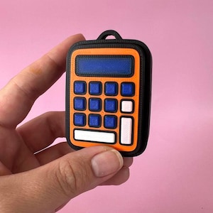 May include: A small, rectangular calculator-shaped object with a black outer frame, an orange faceplate, and a blue display screen. It features nine blue square buttons, two white rectangular buttons, and one white square button, with a small loop at the top.