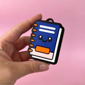 May include: A blue notebook-shaped charm with a black border, featuring a smiling face with two black eyes and a curved mouth. The design includes orange spiral binding, a white horizontal line, and a rectangular orange accent on a light pink base.