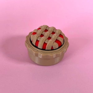 Op de afbeelding: Een kleine, ronde, beige container met een geschulpte rand, voorzien van een deksel dat is ontworpen als een taart met een rasterpatroon. Het deksel heeft een beige rasterpatroon over een felrood cirkelvormig midden, lijkend op een taart.