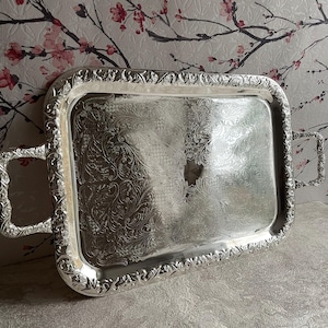 May include: A rectangular silver-coloured serving tray featuring ornate embossed floral and scrollwork patterns along the raised border and handles. The central surface of the tray displays a detailed, etched filigree design with a polished finish.
