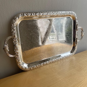 May include: A rectangular silver-toned serving tray featuring an ornate, embossed floral border and matching handles. The central surface displays a detailed, etched scrollwork pattern that reflects light across its polished, metallic finish.