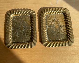 May include: Two rectangular brass trays featuring a central engraved pattern of radiating lines and leaf motifs. Each tray has a raised, scalloped, and twisted edge design that frames the flat, detailed center section of the metallic surface.