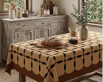 Op de afbeelding: Een rechthoekig tafelkleed met een herhalend geometrisch patroon van in elkaar grijpende cirkels in tinten beige, crème en donkerbruin. De stof ligt over een houten tafel met daarop een brood op een houten plank, een glazen karaf en een vaas.