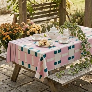 May include: A rectangular tablecloth featuring a geometric pattern of pink, white, and dark teal blocks. The table is set with a white porcelain teapot, two matching teacups on saucers, a small creamer, and a plate of scones on a wooden picnic table.