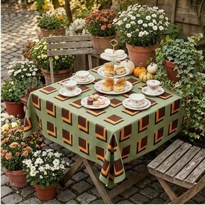 May include: A square tablecloth with a sage green base and a repeating geometric pattern of brown and orange squares. The table is set with white ceramic teacups, saucers, and a tiered serving stand holding baked goods, surrounded by potted flowers.