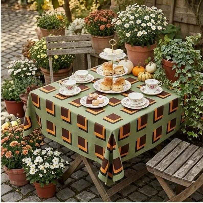 May include: A square tablecloth with a sage green base and a repeating geometric pattern of brown and orange squares. The table is set with white ceramic teacups, saucers, and a tiered serving stand holding baked goods, surrounded by potted flowers.