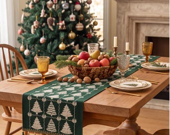 Op de afbeelding: Een donkergroene tafelloper met een wit ruitpatroon, kerstboommotieven en decoratieve kwastjes aan de uiteinden. De loper ligt op een houten tafel gedekt met borden, amberkleurige glazen en een schaal met fruit.