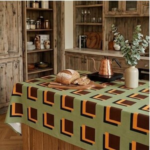 May include: A rectangular tablecloth featuring a repeating geometric pattern of brown, orange, and black squares on a sage green background. The fabric drapes over a wooden kitchen island, providing a flat surface for a loaf of bread and a kettle.