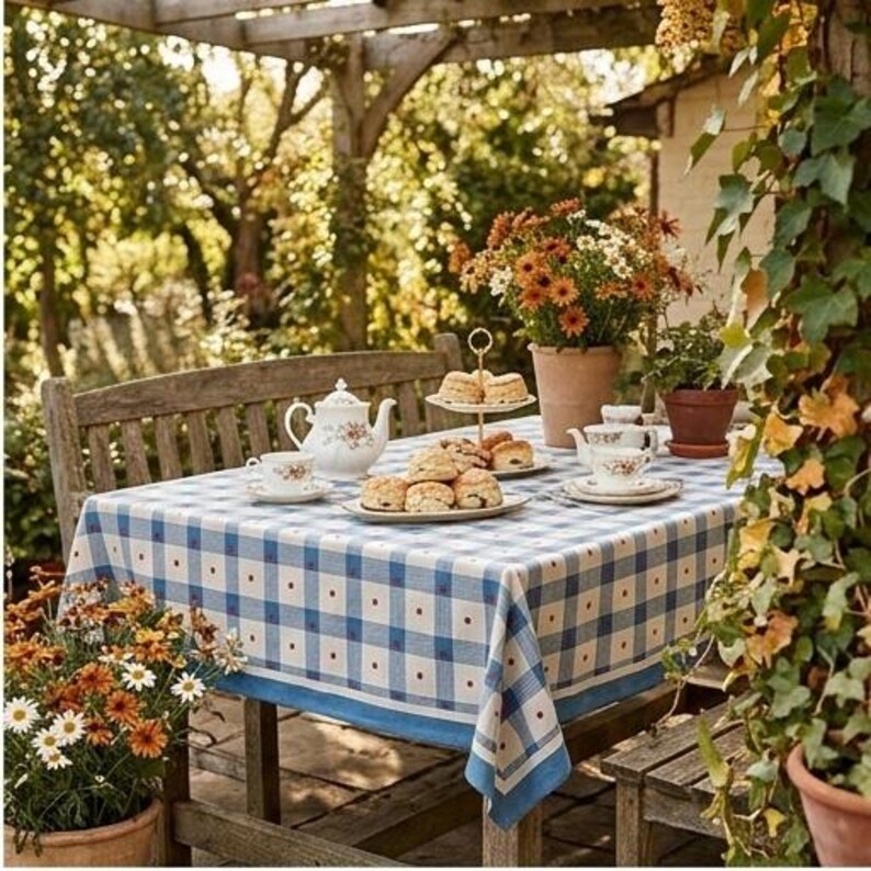 May include: A blue and white checkered tablecloth with small red dots, featuring a white porcelain teapot with floral patterns, matching teacups, and a two-tiered serving stand holding several baked scones on a wooden outdoor table.