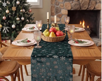 Op de afbeelding: Een donkergroene fluwelen tafelloper met een metallic zilveren sneeuwvlokpatroon op een houten eettafel. De tafel is gedekt met witte borden, glazen met een textuur en een rieten mand gevuld met peren en granaatappels.