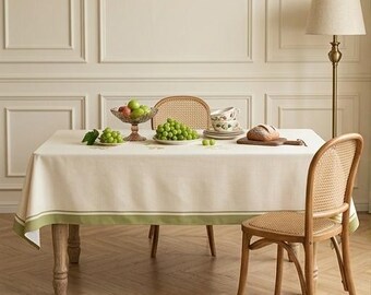 Op de afbeelding: Een rechthoekig gebroken wit tafelkleed met een brede saliegroene rand aan de onderkant. Het tafelkleed ligt over een houten tafel met een glazen fruitschaal, een bord met groene druiven, een stapel bloemmotief kommen en een brood.