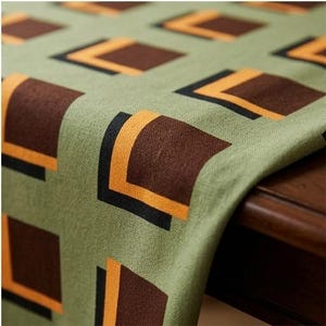 May include: A sage green table runner featuring a repeating geometric pattern of dark brown squares layered with smaller, bright orange and black L-shaped accents. The fabric has a visible woven texture and drapes over the edge of a wooden surface.