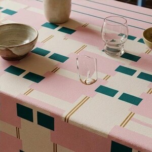 May include: A tablecloth featuring a geometric pattern of pink and cream rectangles, dark teal squares, and thin gold lines. On the surface are a beige ceramic bowl, a clear glass tumbler, a small clear glass vessel, and a small beige bowl.