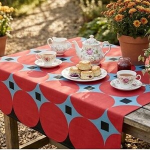 May include: A rectangular tablecloth featuring a repeating pattern of large red circles on a light blue background with small black squares between the circles. The tablecloth is draped over a wooden table set with a floral teapot, cups, and scones.