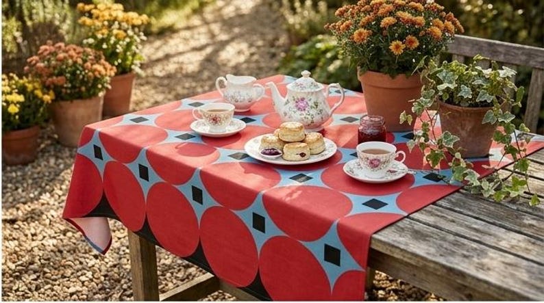 May include: A rectangular tablecloth featuring a repeating pattern of large red circles on a light blue background with small black squares between the circles. The tablecloth is draped over a wooden table set with a floral teapot, cups, and scones.