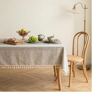 May include: A rectangular light gray tablecloth with a subtle woven texture and white fringe trim along the edges. The tablecloth is draped over a wooden table, featuring a striped pattern, and is paired with a light wood chair with a cane seat.