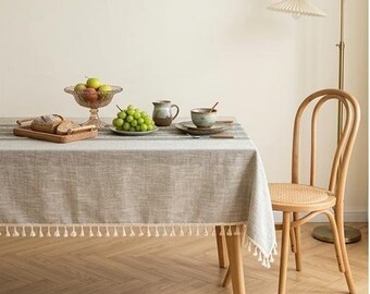 Op de afbeelding: Een rechthoekig lichtgrijs tafelkleed met een subtiele geweven textuur en witte franjes langs de randen. Het tafelkleed ligt over een houten tafel, heeft een gestreept patroon en wordt gecombineerd met een lichthouten stoel met rieten zitting.
