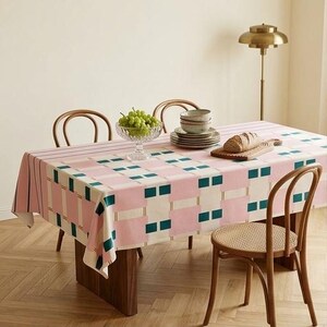 May include: A rectangular tablecloth featuring a geometric pattern of pink, cream, and dark teal squares and stripes. The table is set with a glass bowl of green grapes, a stack of ceramic bowls, and a loaf of bread on a wooden cutting board.