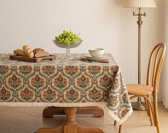 Op de afbeelding: Een rechthoekige houten eettafel bedekt met een crèmekleurig tafelkleed met een herhalend bloemrijk damastpatroon in roestoranje en saliegroen, afgewerkt met een franjerand, vergezeld door een houten stoel en een vloerlamp.