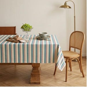 May include: A rectangular tablecloth featuring vertical stripes in shades of teal, cream, and thin dark red lines. The fabric covers a wooden table, which is set with a glass bowl of green grapes, a small wooden cutting board, and a ceramic bowl.