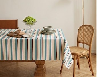 Op de afbeelding: Een rechthoekig tafelkleed met verticale strepen in tinten blauwgroen, crème en dunne donkerrode lijnen. De stof bedekt een houten tafel, die is gedekt met een glazen schaal met groene druiven, een kleine houten snijplank en een keramische kom.