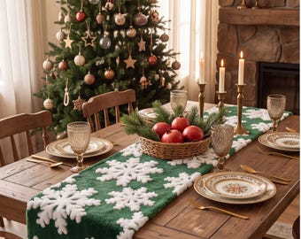 Op de afbeelding: Een groene tafelloper met een wit sneeuwvlokpatroon op een houten eettafel, gedekt met borden, goudkleurig bestek, glazen met textuur en een rieten mand met rode granaatappels en groenblijvende takken.