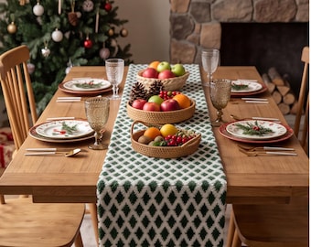 Op de afbeelding: Een houten eettafel gedekt voor een maaltijd met een witte en groene tafelloper met ruitpatroon. De tafel is voorzien van witte borden met kerstmotieven, goudkleurig bestek, glazen met textuur en twee ronde gevlochten manden met fruit.