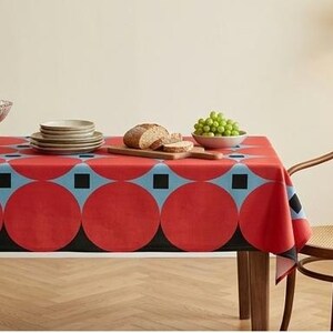 May include: A rectangular tablecloth featuring a bold geometric pattern of large red circles, light blue accents, and small black squares. The fabric covers a dining table set with a glass fruit bowl, a stack of plates, sliced bread, and green grapes.