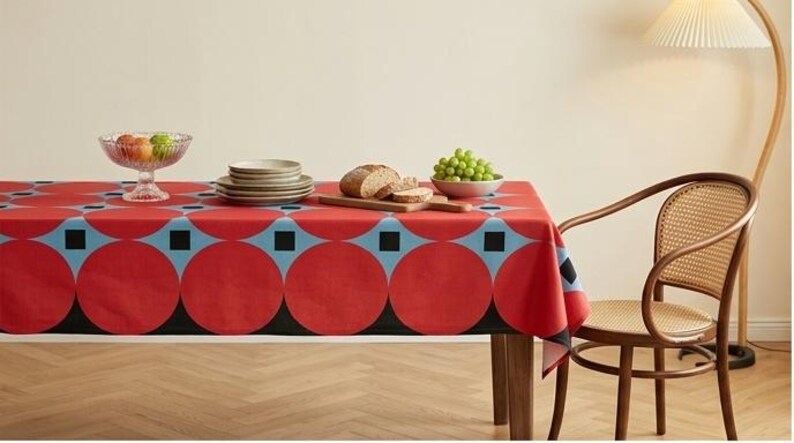 May include: A rectangular tablecloth featuring a bold geometric pattern of large red circles, light blue accents, and small black squares. The fabric covers a dining table set with a glass fruit bowl, a stack of plates, sliced bread, and green grapes.