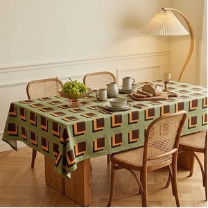 May include: A rectangular tablecloth featuring a repeating geometric pattern of brown and orange squares on a sage green background. The tablecloth is draped over a wooden dining table surrounded by four wooden chairs with woven cane backrests.