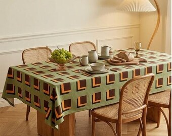 Op de afbeelding: Een rechthoekig tafelkleed met een herhalend geometrisch patroon van bruine en oranje vierkanten op een saliegroene achtergrond. Het tafelkleed ligt over een houten eettafel, omringd door vier houten stoelen met rugleuningen van rotan vlechtwerk.