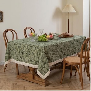 May include: A rectangular tablecloth featuring a dark green botanical leaf pattern with a cream-colored fringe trim along the edges, draped over a wooden dining table with four wooden chairs and a floor lamp in the background.