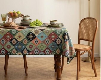 Op de afbeelding: Een rechthoekige eettafel bedekt met een veelkleurig tafelkleed met een herhalend geometrisch ruitpatroon in tinten groen, blauw, rood en crème. Een houten stoel met een rugleuning en zitting van gevlochten riet staat naast de tafel.
