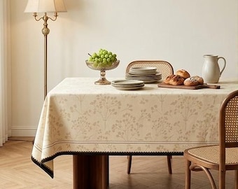 May include: A rectangular tablecloth featuring a light beige fabric with a repeating tan botanical floral pattern. The edges are finished with a dark brown border and a delicate scalloped trim. The cloth covers a wooden table set with plates.