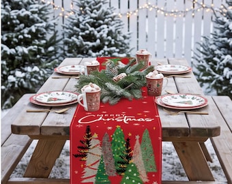 Op de afbeelding: Een rode tafelloper met de witte tekst Merry Christmas en een grafische print van diverse gestileerde kerstbomen in groen, goud en wit, geaccentueerd met kleine sneeuwvlokken en sterren, gedrapeerd over een houten picknicktafel.