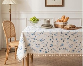 Op de afbeelding: Een rechthoekig tafelkleed van crèmekleurige stof met een delicaat blauw bloemenpatroon en een decoratieve franje langs de randen. Het tafelkleed ligt over een houten tafel en vormt een neutrale basis voor het dekken van de tafel.