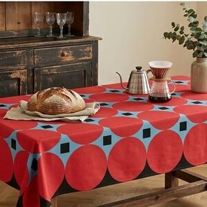 May include: A rectangular tablecloth featuring a repeating pattern of large red circles and small black squares on a light blue background. The fabric drapes over a wooden table, which holds a loaf of bread, a metal kettle, and a glass coffee dripper.