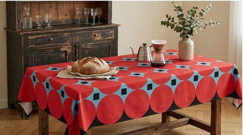 May include: A rectangular tablecloth featuring a repeating pattern of large red circles and small black squares on a light blue background. The fabric drapes over a wooden table, which holds a loaf of bread, a metal kettle, and a glass coffee dripper.