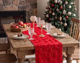 May include: A red velvet table runner with a quilted diamond pattern and gold stitching, placed on a wooden dining table. The table is set with two gold crystal candle holders, clear textured glassware, and a woven basket of fruit.