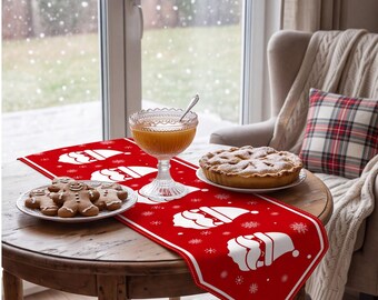 Op de afbeelding: Een rode tafelloper met een herhalend wit patroon van kerstmangezichten en sneeuwvlokken. De loper ligt op een ronde houten tafel met een bord peperkoekmannetjes, een glazen kom met oranje vloeistof en een gebakken taart.