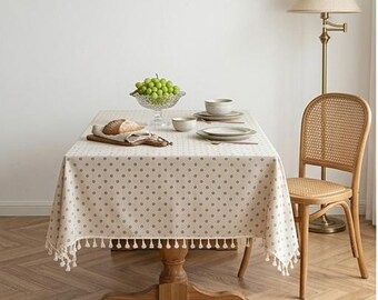 Op de afbeelding: Een rechthoekig beige tafelkleed met een herhalend donkergrijs stippenpatroon en decoratieve franjes langs de randen. Het tafelkleed ligt over een houten eettafel, die is gedekt met een glazen schaal met groene druiven en borden.