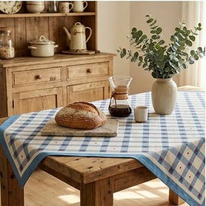 May include: A rectangular tablecloth featuring a blue and white plaid pattern with small red dots, covering a wooden table. On the table sits a round loaf of bread on a stone board, a glass coffee carafe with a brown leather collar, and a vase of eucalyptus.