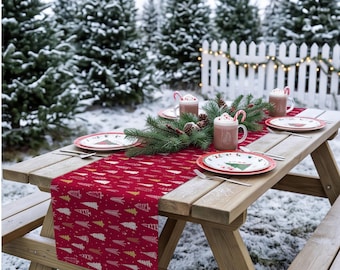 Op de afbeelding: Een rode tafelloper met een herhalend patroon van witte, gouden en donkerrode gestileerde kerstbomen. De loper ligt op een houten picknicktafel gedekt met witte borden, mokken met warme chocolademelk en takken van dennenbomen.