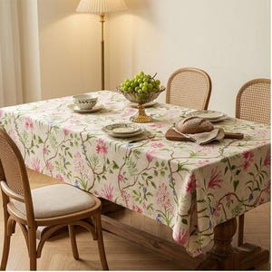 Peut inclure: Une nappe rectangulaire au fond crème ornée d'un motif botanique répétitif de fleurs, de vignes et de feuilles roses, violettes et vertes. Le tissu recouvre une table en bois dressée avec des assiettes, un bol, un pain et des raisins.