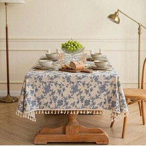 Peut inclure: Une nappe rectangulaire à fond crème avec un motif floral bleu et une bordure à franges décoratives. La nappe recouvre une table à manger en bois disposée avec des bols en céramique, un bol en verre de raisins et du pain.
