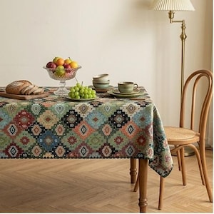 May include: A rectangular tablecloth featuring a repeating geometric diamond pattern in shades of green, red, blue, and orange. The fabric covers a wooden table, which holds a glass fruit bowl, a plate of green grapes, and ceramic tea cups.