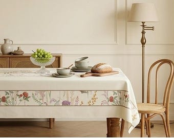 Op de afbeelding: Een rechthoekig, crèmekleurig tafelkleed met een decoratieve rand van kleurrijke wilde bloemen en grassen. De stof ligt over een eettafel, die is gedekt met een kristallen fruitschaal gevuld met groene druiven, keramische kommen en brood.