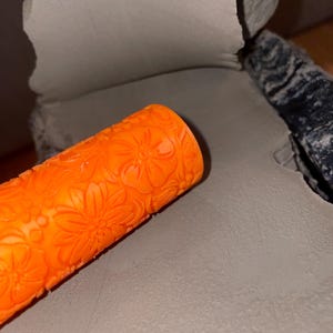 Könnte beinhalten: Eine leuchtend orangefarbene, zylindrische Tonwalze mit erhabenem Blumenmuster. Die Walze liegt auf einer flachen, glatten Platte aus hellgrauem Ton und zeigt die Textur, die beim Eindrücken in die Oberfläche des formbaren Materials entsteht.