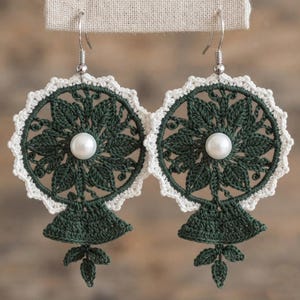 May include: A pair of round crochet earrings featuring a dark forest green floral pattern with a central white pearl bead. The circular design is bordered by a scalloped white edge and includes a small dangling leaf motif at the bottom.